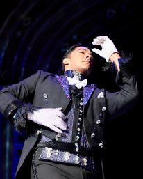 Stage performer in a sparkling black tuxedo with jeweled details and white gloves striking a dramatic pose on a theater stage under bright stage lighting