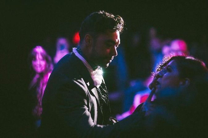 Moody nightclub scene: a man in a dark jacket leans toward a woman on a crowded dance floor bathed in purple and blue neon lights.