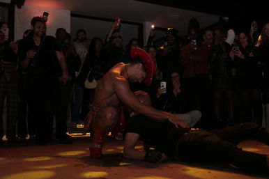 Male entertainer in a red sequined cowboy hat and costume crouches over a person on the dance floor while a cheering nightclub crowd records the playful performance on phones under dim spotlights.
