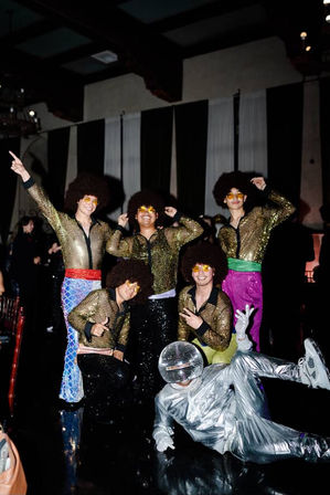 Disco-themed group posing on an indoor dance floor: five people in gold sequin jackets, colorful bell-bottoms, large afro wigs and yellow star sunglasses, plus one person reclining in a silver mirror-ball helmet suit at a party venue.