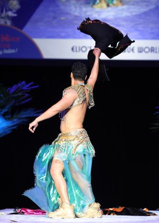 Stage performer tossing a black hat while wearing a turquoise shimmering skirt with a gold-coin hip scarf, beaded shoulder harness and gold sneakers during a lively live show.