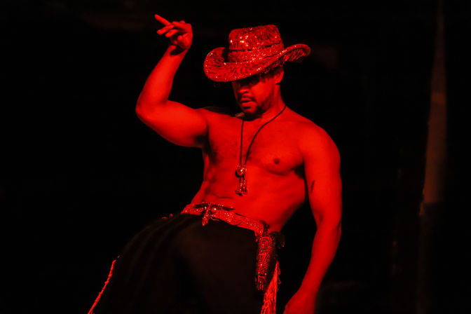 Shirtless male dancer in a glittery cowboy hat and tasseled belt striking a dramatic pose on a dark stage bathed in red light.