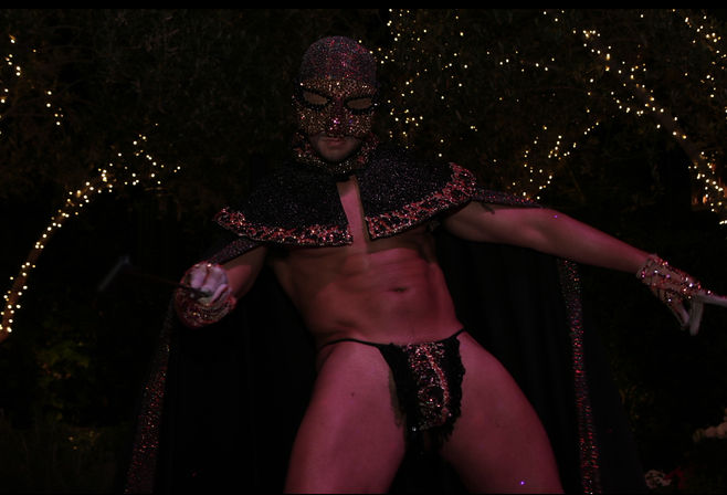 Masked male performer in a glittering beaded mask, cape and embellished thong striking a pose under twinkling string lights at an outdoor nighttime garden party