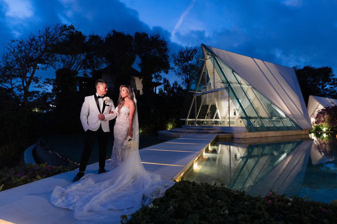 Bride in lace gown and groom in white tuxedo on an illuminated walkway beside a modern glass chapel reflected in a serene pool at dusk, romantic destination wedding scene.