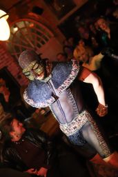 Masked performer in a glittering beaded cape, sequined mask and sheer bodysuit struts through a crowded, dimly lit nightclub with exposed brick and arched window.