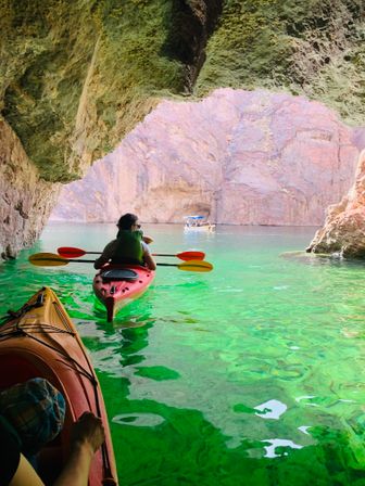 Kayaker paddling from a shaded rock cave into emerald-green canyon waters with sunlit pink-red cliffs in the background