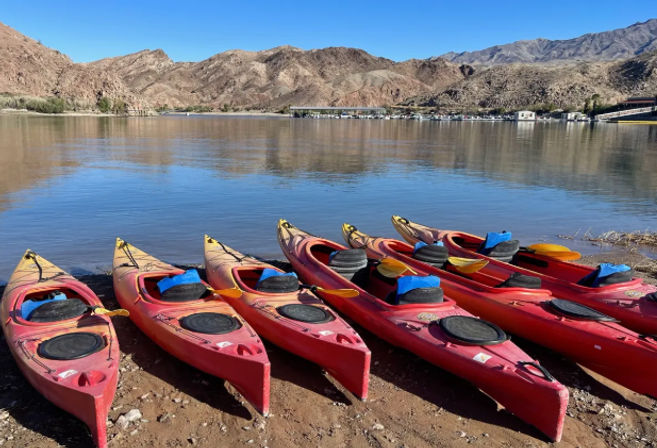 Bright red kayaks lined up on a sandy lakeshore beside a calm desert lake, framed by rocky mountains and a clear blue sky.