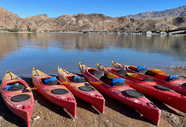 Bright red kayaks lined up on a sandy lakeshore beside a calm desert lake, framed by rocky mountains and a clear blue sky.