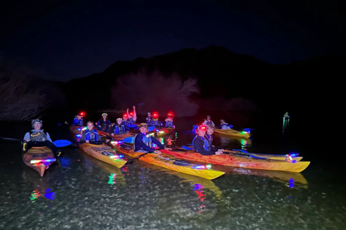 Group of kayakers glowing with colorful LED lights and headlamps, sitting in yellow and orange kayaks on calm water at night with bright reflections and a dark shoreline silhouette