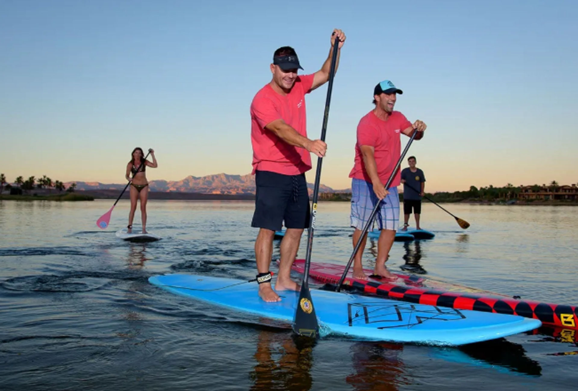 Four stand-up paddleboarders enjoying a calm sunset lake paddle with a mountain backdrop and shoreline homes, two men leading and others following.