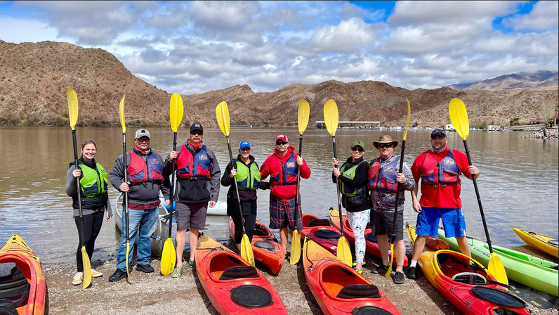 Eight kayakers in life jackets holding yellow paddles beside red kayaks on a desert-lake shoreline with rugged brown mountains under a partly cloudy sky, ready for an outdoor kayaking adventure.