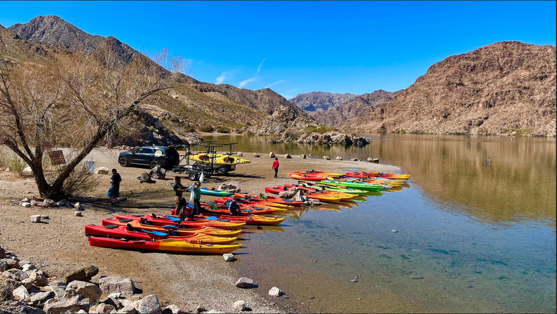 Vibrant red, yellow and green kayaks lined up on a sandy desert lake shore with people preparing to launch, calm blue water and rugged canyon mountains under a bright sky.