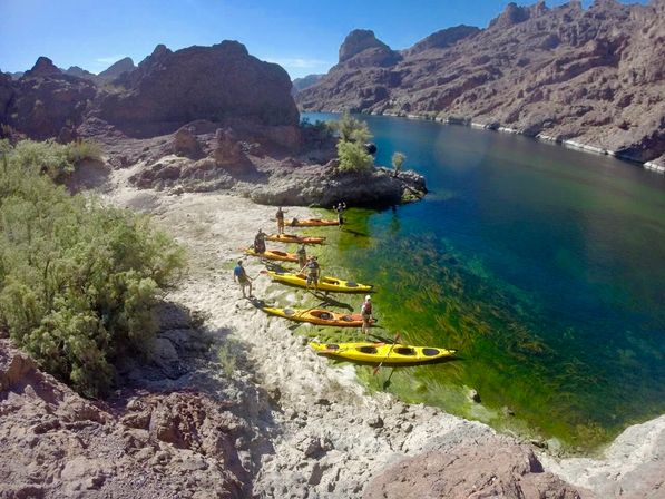 Group of yellow and orange kayaks beached on a sandy cove beside clear blue-green river winding through a rugged desert canyon under a sunny sky