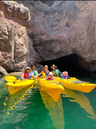 Group of kayakers in bright yellow kayaks and life vests paddling on emerald-green water at the mouth of a sunlit rocky canyon cave