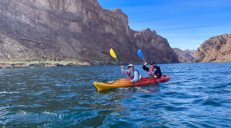 Two kayakers in a yellow-orange tandem kayak paddling on deep-blue river water below towering desert canyon cliffs under a clear blue sky