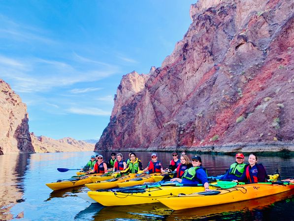 Sunlit group of kayakers in yellow kayaks paddling on a calm canyon lake beneath towering red-rock cliffs and a clear blue sky.