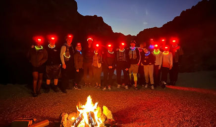 Nighttime desert canyon camp: group of hikers with red headlamps and neon glow necklaces gathered around a roaring campfire under a twilight sky