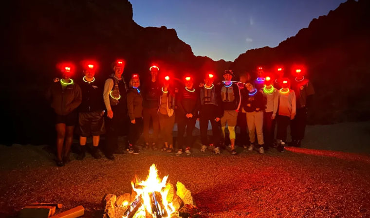 Nighttime desert canyon camp: group of hikers with red headlamps and neon glow necklaces gathered around a roaring campfire under a twilight sky