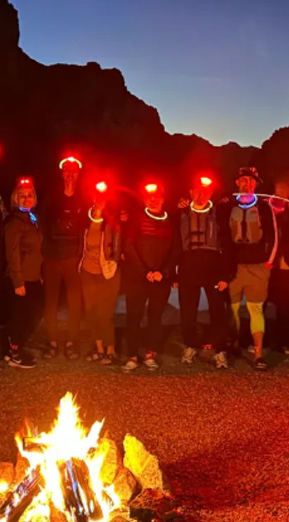 Nighttime desert canyon camp: group of hikers with red headlamps and neon glow necklaces gathered around a roaring campfire under a twilight sky