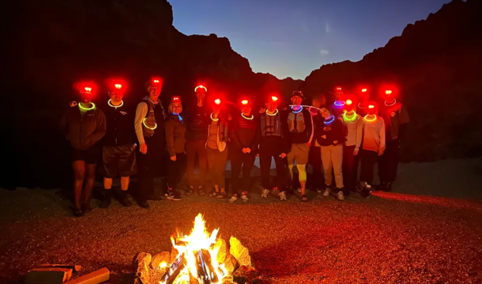 Nighttime desert canyon camp: group of hikers with red headlamps and neon glow necklaces gathered around a roaring campfire under a twilight sky