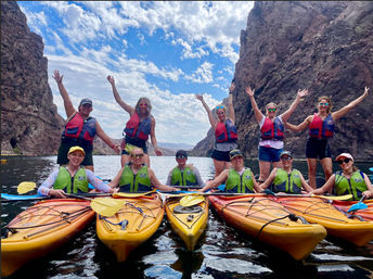 Group of kayakers in colorful life jackets linked together on orange kayaks in a narrow desert canyon, raising hands and smiling on calm water under a blue, partly cloudy sky.