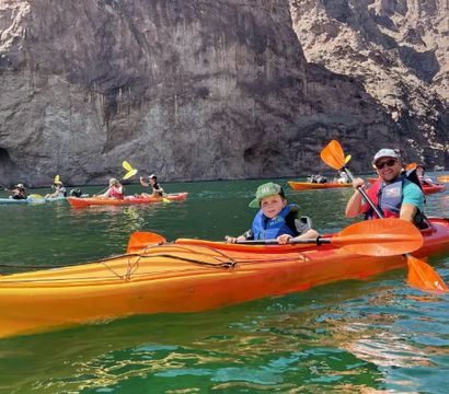 Adult and child paddling an orange tandem kayak on emerald-green water beneath towering rocky canyon walls, with other kayakers nearby