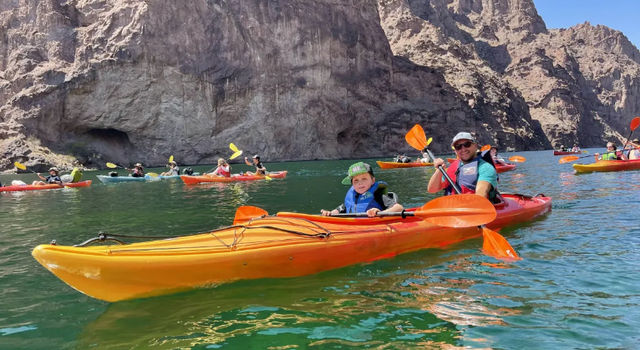 Adult and child paddling an orange tandem kayak on emerald-green water beneath towering rocky canyon walls, with other kayakers nearby