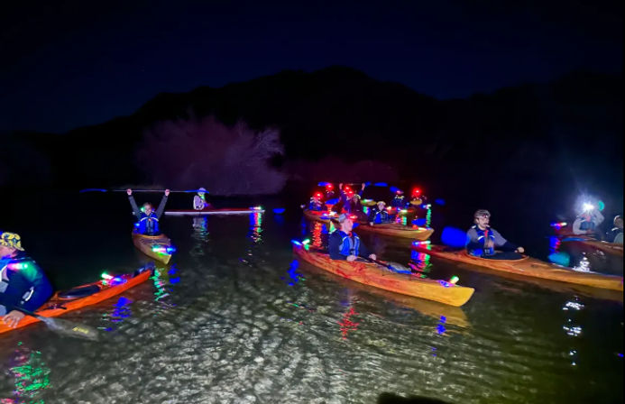 Night kayaking on a calm mountain lake—group of paddlers in life vests in LED-lit kayaks with colorful lights reflecting on the water