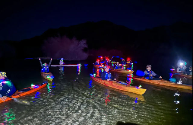 Night kayaking on a calm mountain lake—group of paddlers in life vests in LED-lit kayaks with colorful lights reflecting on the water