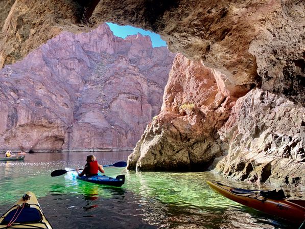 Kayakers paddling from a shaded cave onto clear green river water, framed by towering red-rock canyon cliffs under a bright blue sky.