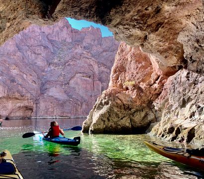Kayakers paddling from a shaded cave onto clear green river water, framed by towering red-rock canyon cliffs under a bright blue sky.