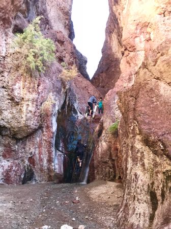 Three hikers climb a rusty metal ladder next to a trickling waterfall in a narrow red‑rock slot canyon — arid desert setting with steep sandstone walls.
