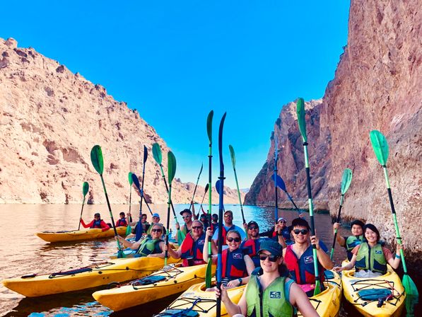 Cheerful group of kayakers in yellow kayaks holding green paddles in a narrow desert canyon with rust-colored cliffs, bright blue sky, and calm water — outdoor kayaking adventure.