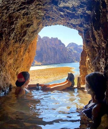 Two people relaxing in a rocky cave pool, gazing out through a cave opening at a sunlit river canyon and sandy riverbank — scenic cave pool river view