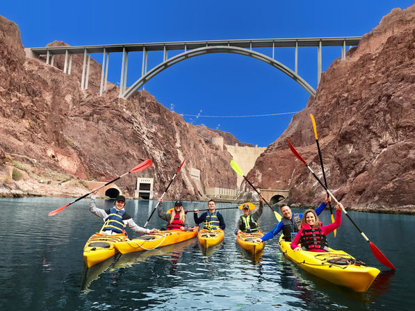 Group of six kayakers in yellow kayaks raising paddles on a calm stretch of the Colorado River beneath the arched bridge and Hoover Dam, framed by red rock canyon walls and a bright blue sky