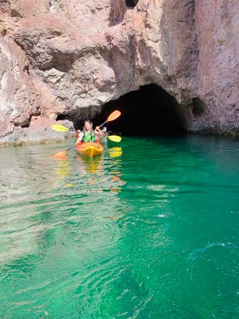 Person paddling an orange kayak toward a dark sea cave under sunlit pinkish rock cliffs, vivid turquoise-green water