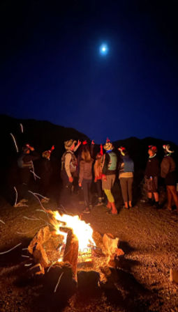 Group gathered around a glowing campfire at a nighttime mountain campsite, pointing up at a bright moon while stargazing
