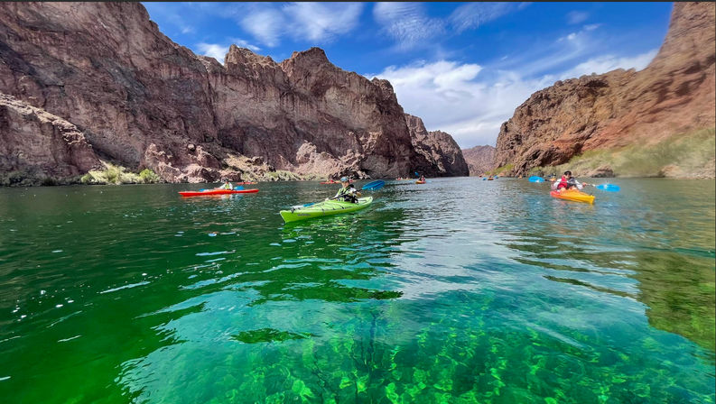 Colorful kayaks and paddlers gliding on emerald-clear river water through towering red-brown canyon cliffs under a bright blue sky