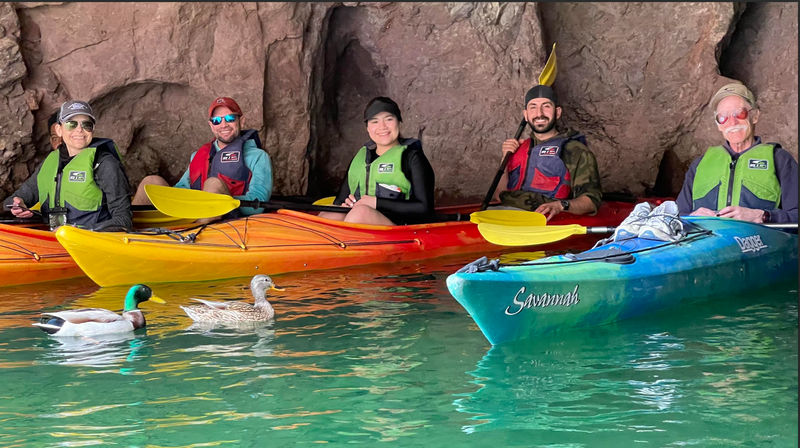 Five kayakers in colorful kayaks inside a rocky sea cave, paddling on clear emerald water while two mallard ducks swim nearby.