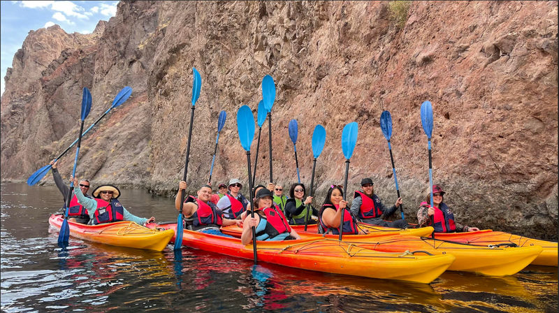 Group of kayakers wearing red life jackets in yellow and orange kayaks raising blue paddles while floating beside a steep rocky canyon wall on calm water.