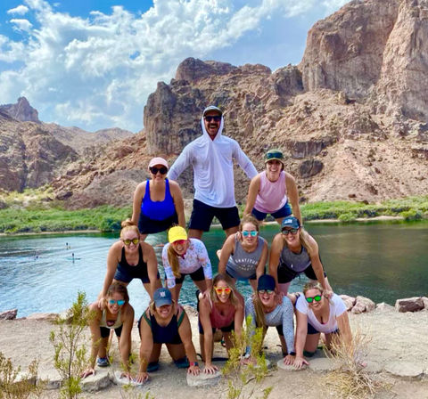 Friends in activewear forming a three-tier human pyramid on a sandy desert canyon riverbank with turquoise water, paddleboarders in the distance and dramatic rocky cliffs under a bright blue sky