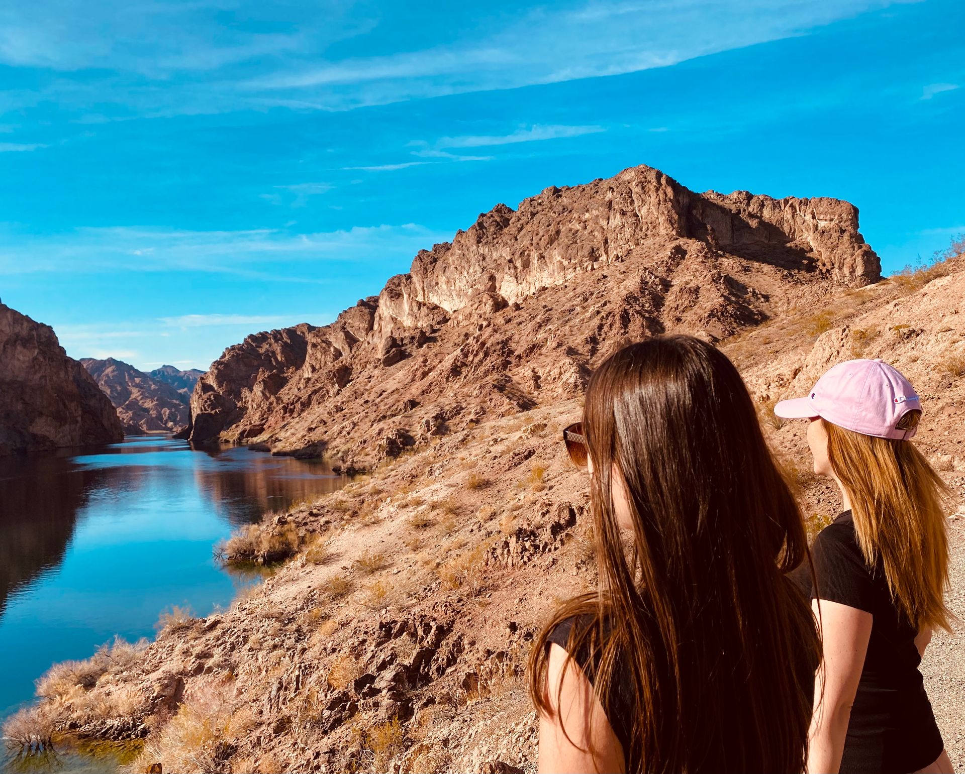 Two hikers overlooking a winding blue river cutting through a rugged desert canyon under a bright blue sky