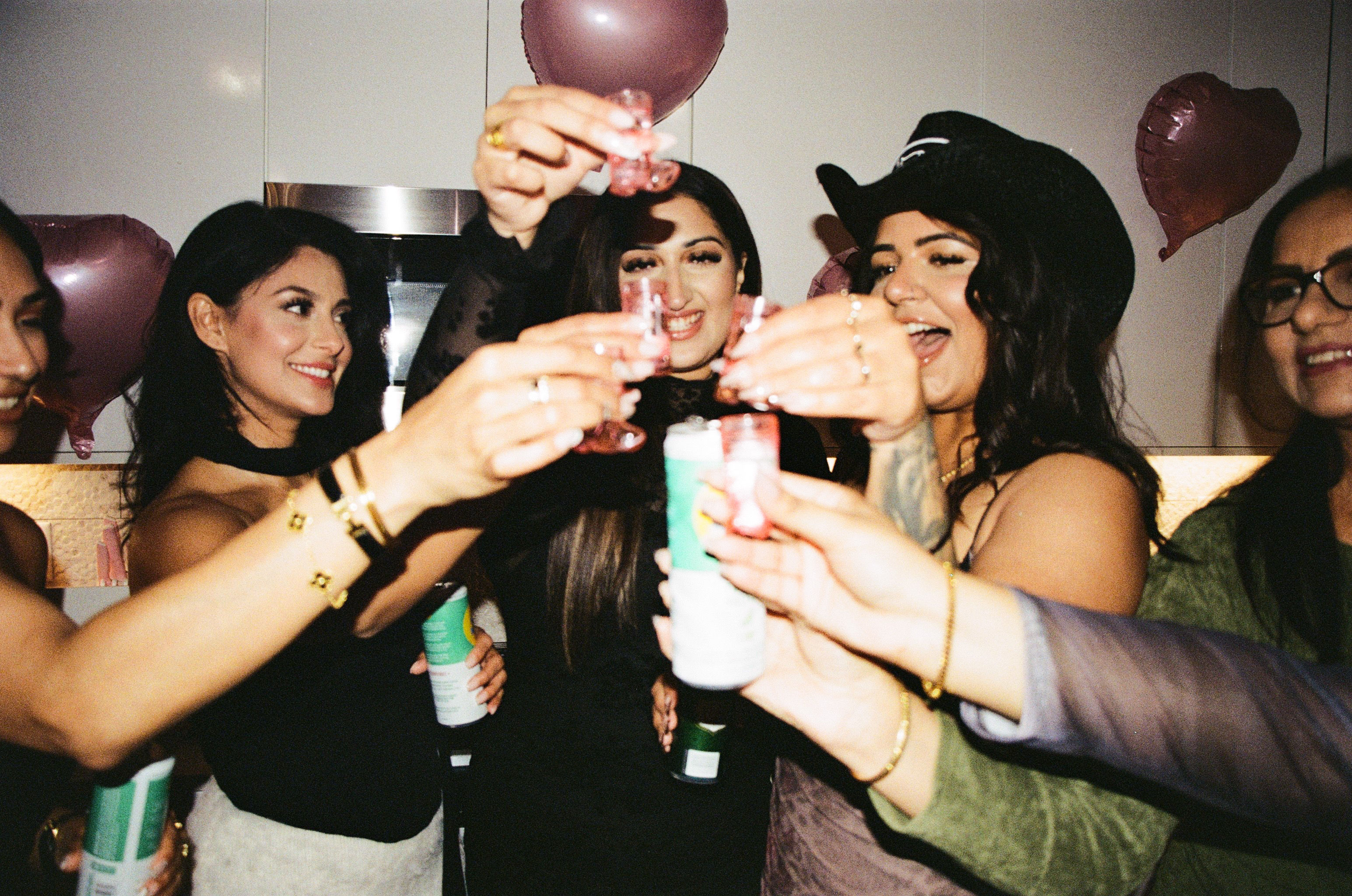 Friends clinking shot glasses and seltzer cans at an indoor party, smiling and cheering under pink heart balloons.