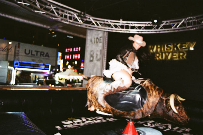 Person in cowboy hat and boots riding a mechanical bull under truss lights with neon signs and a blurred nighttime crowd in a lively outdoor bar scene.