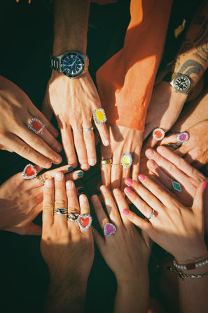 Sunlit overhead close-up of multiple hands arranged in a circle wearing colorful heart and oval gemstone rings, watches, bracelets and painted nails.