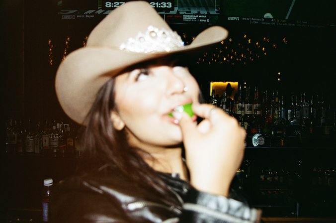 Blurred close-up of a person wearing a cowboy hat and leather jacket popping a lime into their mouth at a dimly lit bar with bottles and TV screens in the background