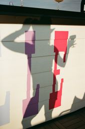 Playful shadow silhouettes of two people cast on a cream wall decorated with purple and red geometric blocks, one tipping a hat while the other holds a drink, with wine glasses on a rooftop railing above.