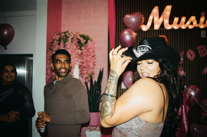 Indoor party scene: tattooed woman wearing a black cowboy hat strikes a playful pose in front of pink balloons and floral backdrop while friends smile and chat nearby.