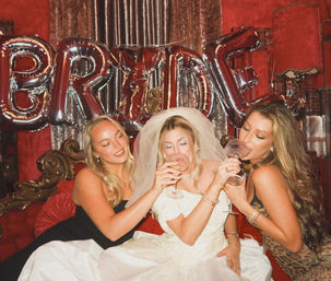 Bride-to-be in a veil and white gown sipping cocktails with two friends on a red velvet vintage sofa beneath metallic 'BRIDE' balloons at an indoor bachelorette party