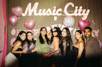 Birthday party photo of friends posing under an illuminated "Music City" sign with pink heart balloons and a birthday banner
